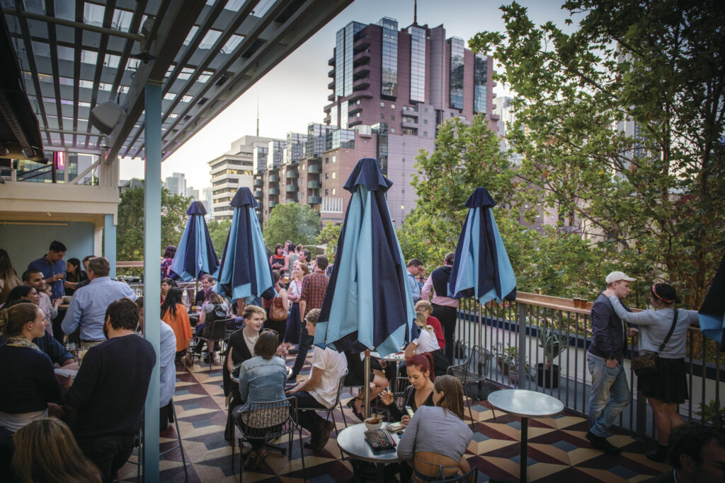 Groups of people sit at tables around closed blue umbrellas at a rooftop bar in Melbourne Australia. The tiled floor is patterned. There is a view of the city behind the people.