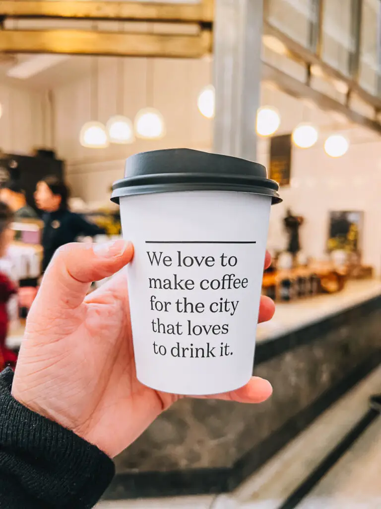 A hand holding a disposable coffee cup with the phrase "We love to make coffee for the city that loves to drink it" in the foreground, with a blurred coffee shop scene in the background.