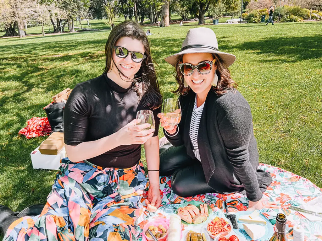 Two women (the author on the left and her friend) enjoying a picnic, toasting with wine glasses in a grassy park. They are seated on a colourful picnic blanket with various foods spread out in front of them. A Melbourne Mystery Picnic is a great activity to share with friends, family or your other half.