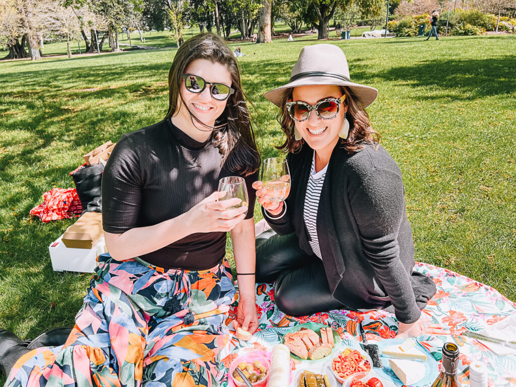 Two women (the author on the left and her friend) enjoying a picnic, toasting with wine glasses in a grassy park. They are seated on a colourful picnic blanket with various foods spread out in front of them. A Melbourne Mystery Picnic is a great activity to share with friends, family or your other half.