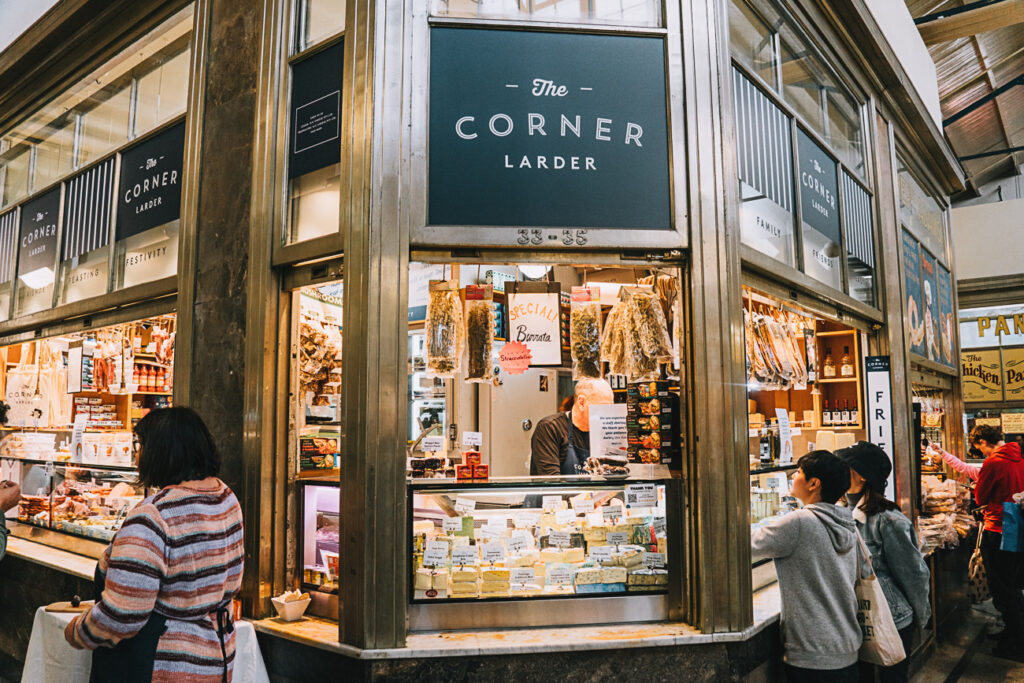 Exterior view of 'The Corner Larder' shopfront in Queen Victoria Market in Melbourne, offering meats and cheeses, with customers at the counter and others walking by. The Queen Victoria Market, where our AmazingCo mystery picnic Melbourne began.