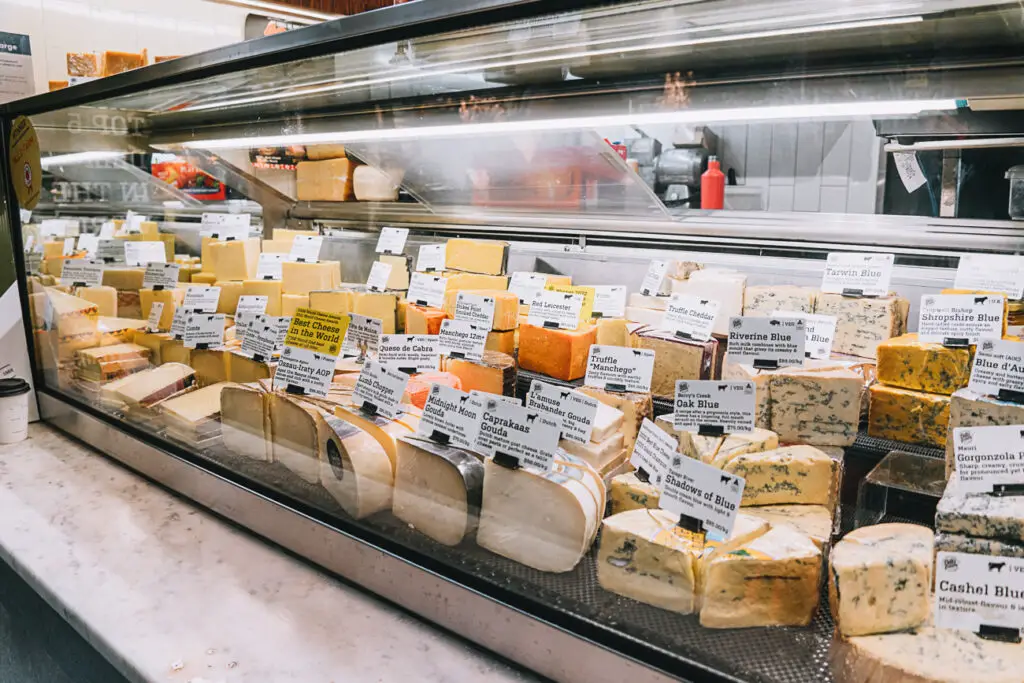 A cheese display case filled with a wide variety of cheeses, each labeled with names and prices, inside Melbourne's iconic Queen Victoria Market. So many of the amazing cheeses on display at Queen Vic Market.