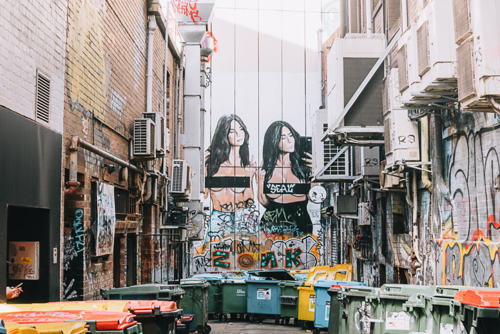 A narrow alley with graffiti artwork depicting Kim Kardashian and Emily Ratajkowski on a wall, surrounded by various bins and urban fixtures. An AmazingCo Mystery Picnic Melbourne also includes stops to see some of Melbourne's cool street art.