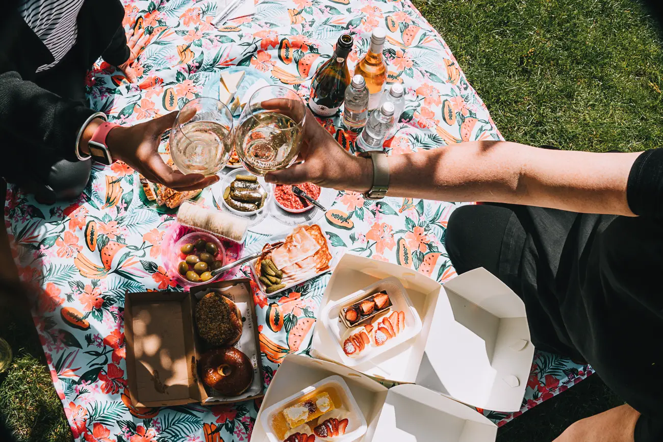 Overhead view of two people's hands toasting with wine glasses over a picnic spread that includes boxes of cakes, dips, and a cheese platter on a floral picnic blanket. The delicious picnic spread we ended up with on our AmazingCo Mystery Picnic Melbourne.