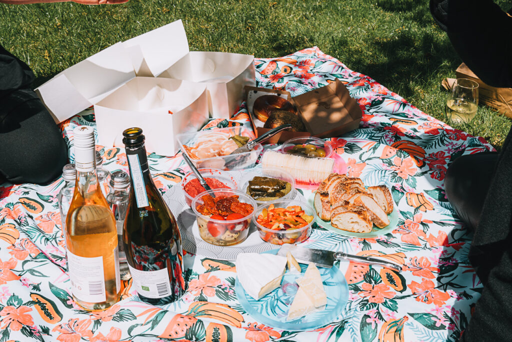 Close-up of a picnic spread on a floral blanket, featuring an assortment of cheeses, bread, olives, and dips, with two bottles of wine and glasses partially visible. The delicious spread we ended up with on our AmazingCo Mystery Picnic Melbourne.