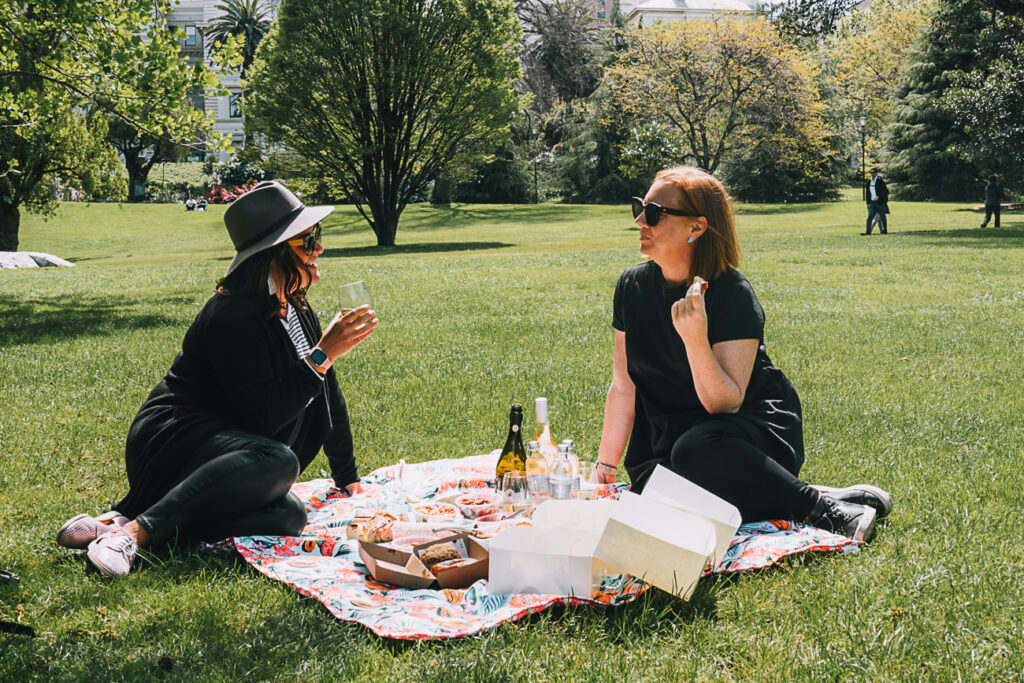 A woman in a hat and sunglasses holds a wine glass, talking to another woman in black attire on a picnic blanket with an array of snacks and open food containers. These are the author's friends, who accompanied her on the mystery picnic. A Melbourne Mystery Picnic is a great activity to share with friends, family or your other half.