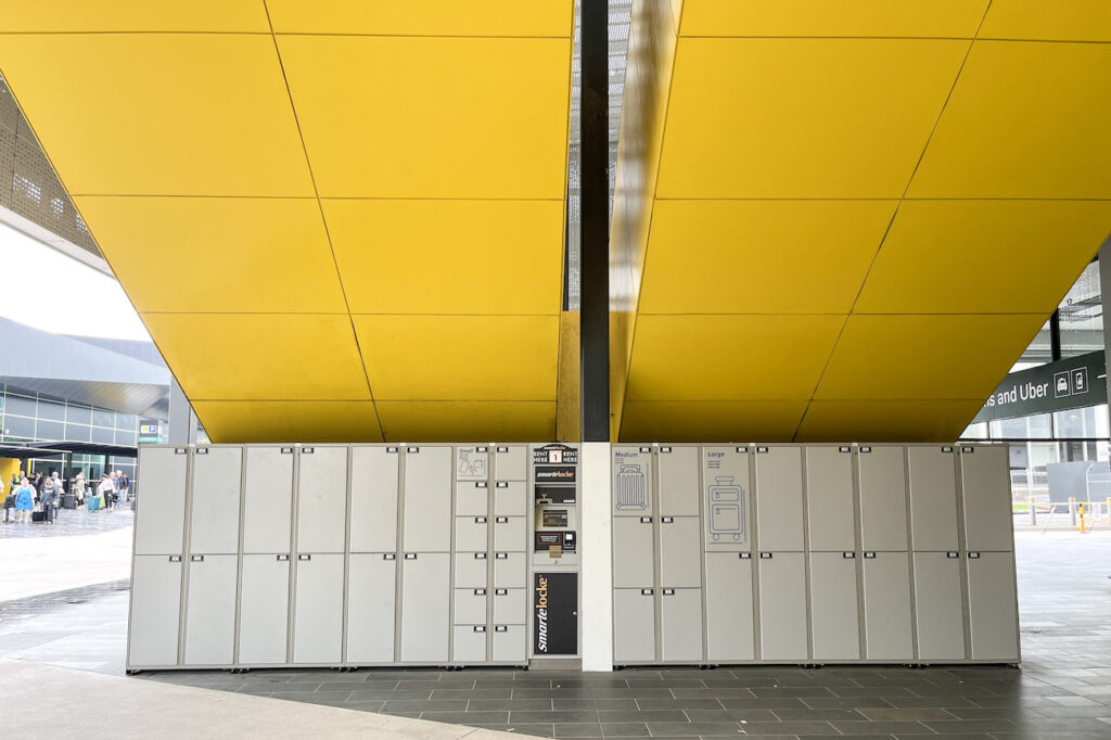 Rows of grey Smartcarte lockers for luggage storage at Melbourne Airport, positioned under bold yellow architectural panels. A central kiosk offers rental options for small, medium, and large suitcases.