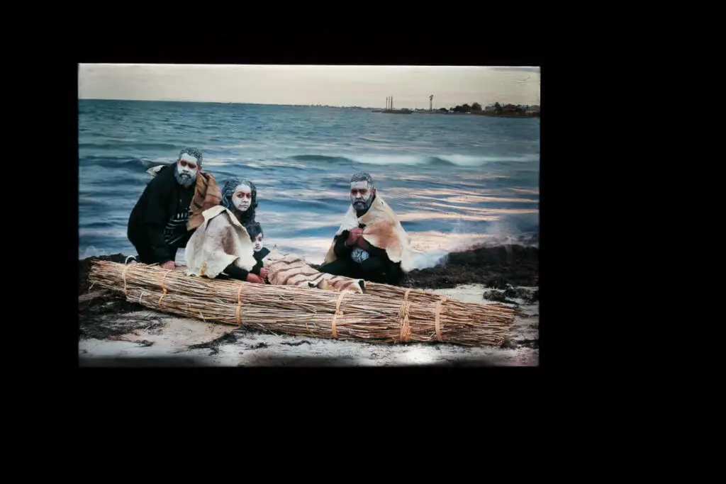 The Ian Potter Centre is known for its Australian art, particularly Indigenous art. A frame from a still movie at an exhibition at the gallery shows three Indigenous people with white painted faces and skin cloaks around their shoulders. They are in a canoe by the water.
