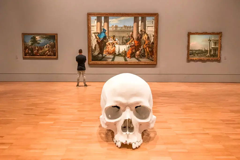The NGV is one of the best museums in Melbourne - they have interesting exhibitions like this one which shows a larger than life human skull in front of three paintings on a wall. A man in a black t-shirt and khaki pants is looking at one of the paintings.