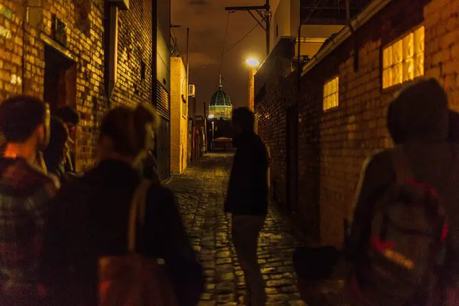 A man leads a ghost tour in Melbourne for a group of people down a dark and shadowy laneway with cobblestones