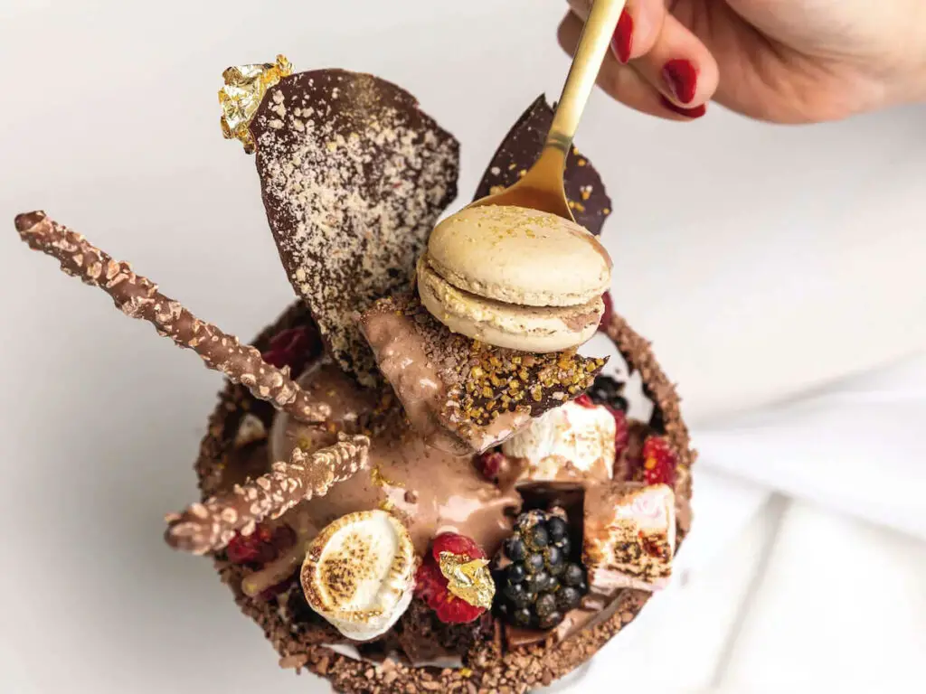 A person dips a gold spoon into a decadent chocolate dessert that is topped with berries, toasted marshmallows and a macaron. One of the best foodie tours in Melbourne is the Melbourne Lanes and Arcades Chocolate and Dessert Walking Tour