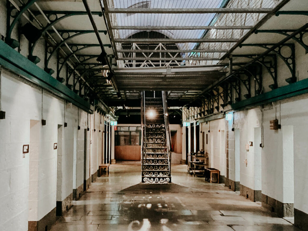 This image shows an indoor corridor in the notorious H Division in Pentridge Prison in Melbourne, Australia, featuring architectural metalwork. The floor appears to be polished, reflecting some of the overhead lighting. The corridor is lined with a series of doors on both sides. The ceiling has exposed metal beams, and there is a walkway with a railing visible on an upper level. Decorative metal spirals adorn the staircase, visible on the right, leading to the upper floor. 