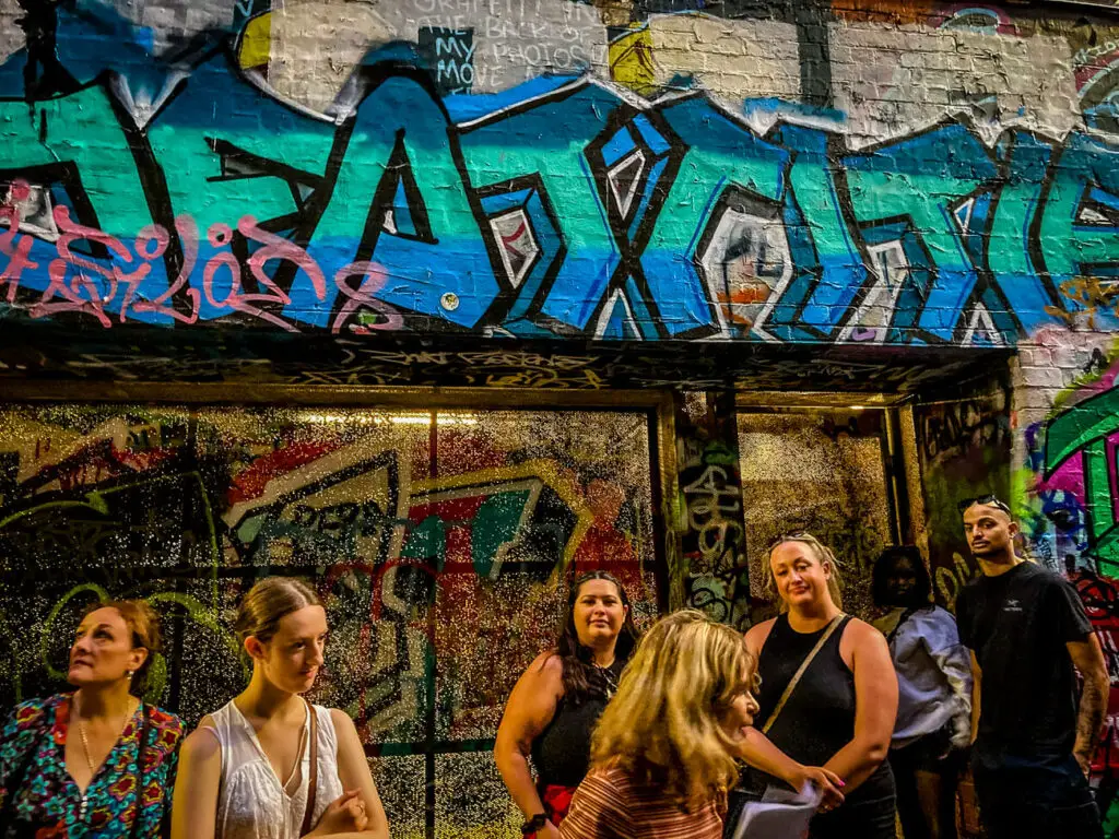 A group of people watch a tour guide leading an Old Melbourne Ghost Tour. They stand in a graffitied alleyway, thought to be a haunted place.