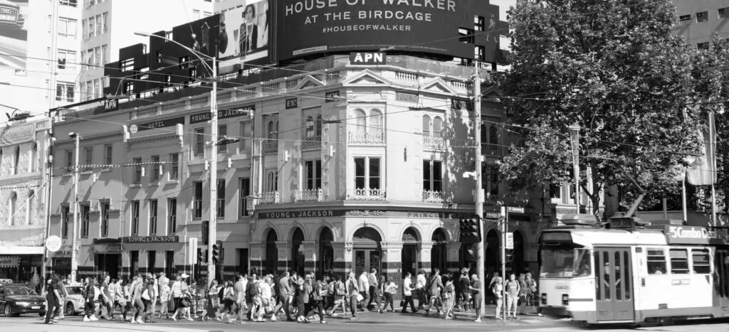 The outside of Young and Jackson Hotel in Melbourne, Australia. There are crowds of people walking past the pub as they cross the street, and a tram driving by - many may not know this is one of Melbourne's haunted pubs