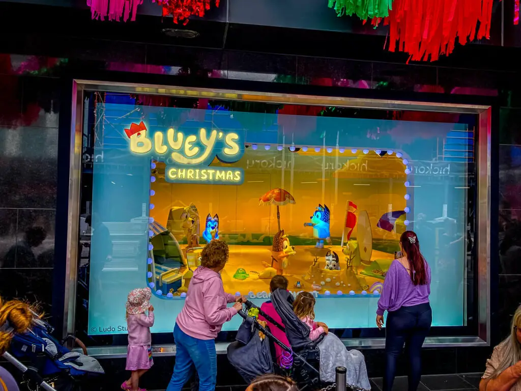 The vibrant Myer Christmas windows display in 2023 showcasing 'Bluey's Christmas' with colourful animated dog characters in a festive scene. Onlookers, including children and adults, some with strollers, are viewing the display. Above the display, bright red and green fringe decorations hang, contributing to the holiday atmosphere.