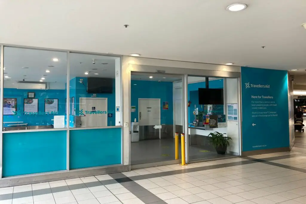 A photograph of the Travellers Aid facility inside Flinders St Station in Melbourne, with a bright turquoise and white colour scheme. The glass doors and windows display the Travellers Aid logo, and there are various informational signs and flyers visible. Travellers Aid offers assistance to travellers, including luggage storage in Melbourne. 