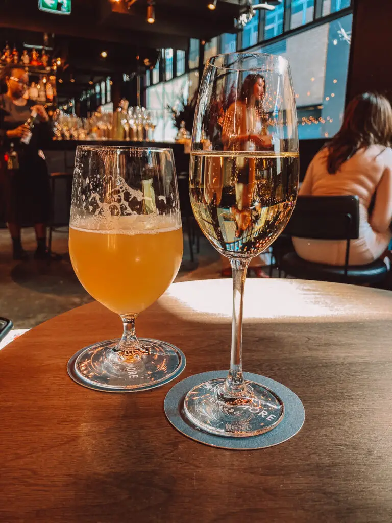 A glass of golden champagne and a cloudy yellow beer on a wooden table. The coasters underneath have "LANCEMORE" printed on them. In the background, a bar with patrons and shelves of bottles. There's a nightly happy hour at the Lancemore Crossley St hotel in Melbourne.