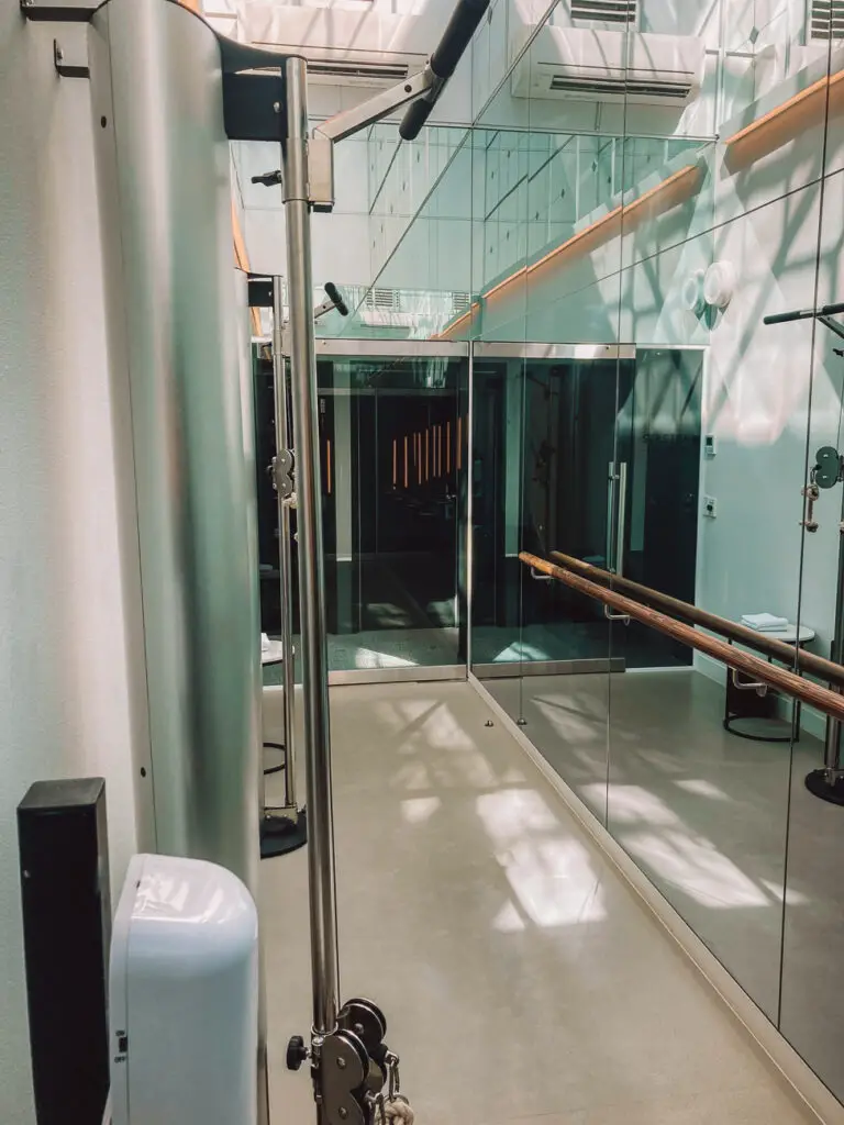 An interior view of a gym with mirrors, a silver pilates reformer machine, and exercise equipment. The room has a clean, modern look with ample natural light. The gym at the Lancemore Crossley St hotel in Melbourne is minimal.