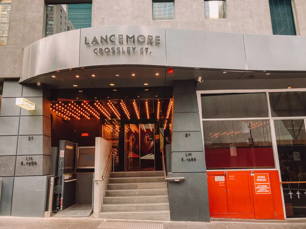 The exterior of the "LANCEMORE CROSSLEY ST." hotel building. The entrance features a metal canopy with lights, two red doors. The Lancemore Crossley St hotel is a great choice for a visit to Melbourne, Australia.